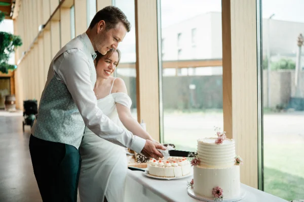 Brautpaar schneidet lachend die Hochzeitstorte an großen Fensterfronten an
