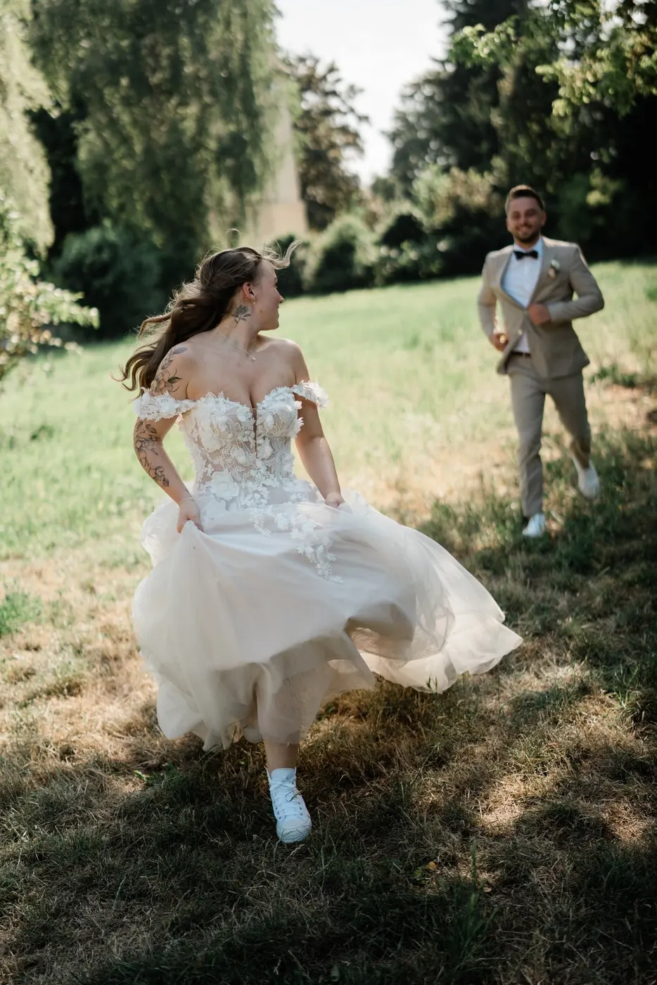 Braut läuft lachend mit gerafftem Kleid über eine Wiese zu ihrem Bräutigam